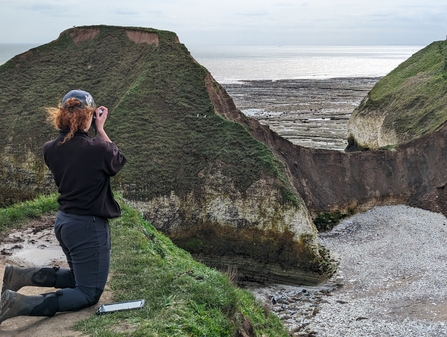 woman kneeling on cliff edge looking through binoculars down onto the beach below where there are grey seals