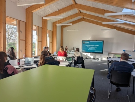 A large green table in the foreground, with students in the mid-ground and a presentation on a blue screen in the background.