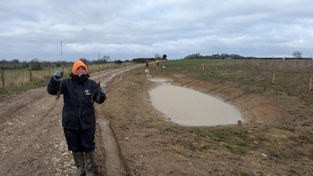 A happy person with two sticks stands to the left of a large, muddy depression with some water in it.