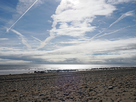 Wide shot of a sandy and shingly beach and a blue sky with clouds and plane trails.