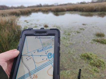 A small handheld device like a phone displays an OS map, held in front of a boggy area of ground