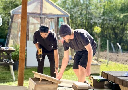 Two men build a wooden structure outdoors on a sunny day. One man uses a power drill while the other inspects the structure. A garden and a greenhouse are visible in the background.
