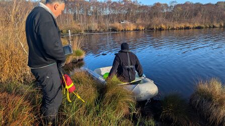 person on a small dingy on the water at the edge. Another man is stood on the bank monitoring.