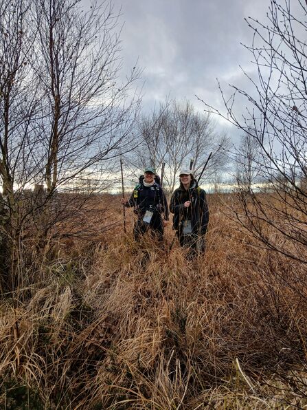 Two people measuring peat depths. They are stood in the middle of a winter field with trees wither side of the frame, facing the camera