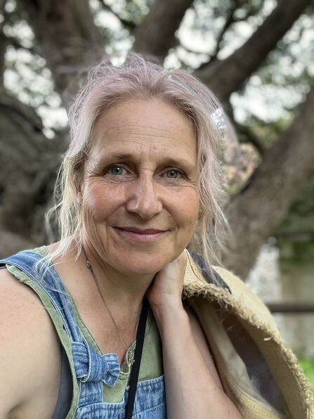 Woman with pink-tinged blonde hair smiles, wearing denim overalls and a green top, resting her hand on a straw hat held on her shoulder, with a blurred tree background.