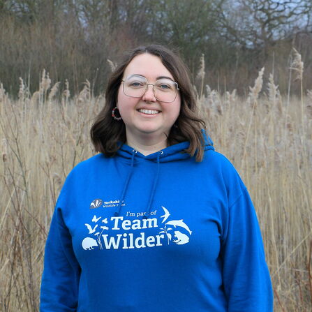 A smiling woman wearing a blue hoodie with "Team Wilder" text, standing in front of tall, dry reeds. She wears glasses, hoop earrings, and a nose ring. The background features a blurry view of trees.