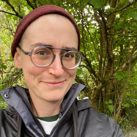 A person with short hair, wearing a burgundy beanie and round-rimmed glasses, smiles at the camera, with a lush green forest background.