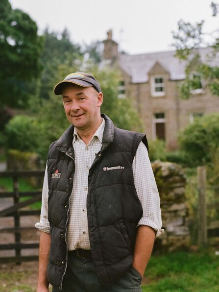 A smiling middle-aged farmer, wearing a baseball cap, a black puffer vest over a checkered shirt, and green pants, stands outdoors with his hands in his pockets in front of a stone house with trees and a wooden fence visible in the background.