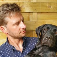 A colour photo showing Tom on the left in a blue shirt looking fondly down at a black dog he's holding 