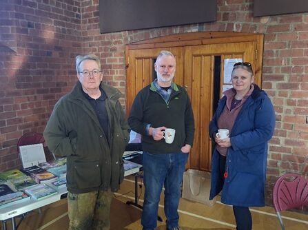 Three adults stand in front of a wooden door and brick wall, with tables of books to their left.