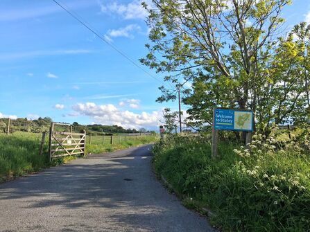 A scenic pathway leads into a green field with a wooden gate on the left and a "Welcome to Stirley" sign with a map on the right, under a bright blue sky with fluffy clouds.