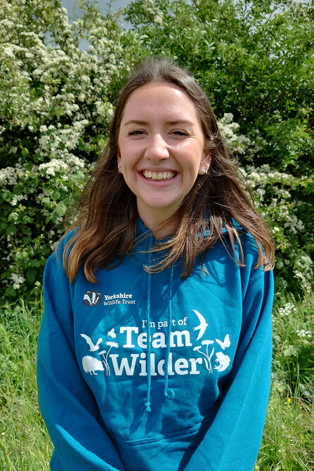 A woman in a teal "Team Wilder" hoodie stands in front of a white-flowered bush.