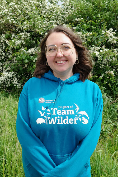 A smiling person wearing glasses and a teal blue hoodie with "Team Wilder" and various wildlife silhouettes printed on it, standing in front of a blooming white bush and tall green grass.