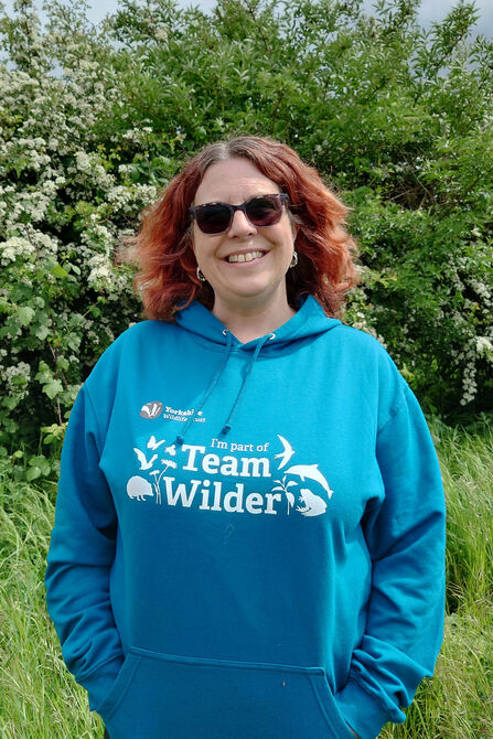 A smiling woman with red hair and sunglasses wears a teal hoodie with "I'm part of Team Wilder" and wildlife graphics, standing outdoors in front of a flowering bush.