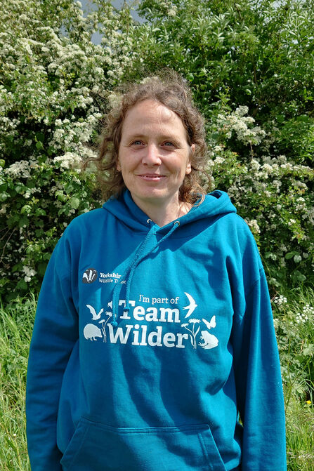 A woman in a teal "Team Wilder" hoodie stands in front of a white-flowered bush.