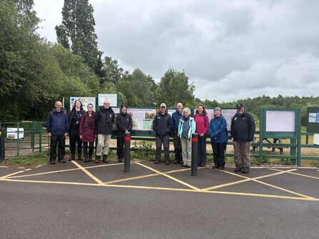 A group of people in outdoor clothing stand in front of interpretive signs at what appears to be the entrance to a nature reserve or park on a cloudy day.