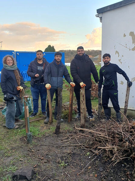 A group of five people stand in a muddy yard outdoors next to a pile of branches and wooden stakes, with a blue utility structure and a weathered building in the background.