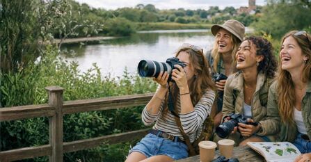 group of friends birdwatching and laughing, one holds binoculars to their eyes and there are some coffee cups and bird id book on the table. Behind them is a nature reserve setting with a wooden fence and a body of water behind it.