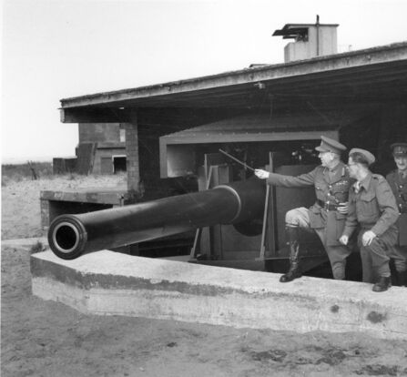 Men in military uniforms stand next to a large coastal artillery gun within a protective concrete emplacement and wooden structure, with one man pointing at the gun.