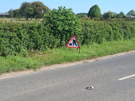 Warning sign depicting a person shoveling dirt next to a roadside hedge.