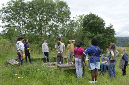 A group of people wearing aprons and safety gloves stand in a grassy outdoor area, listening to a speaker.