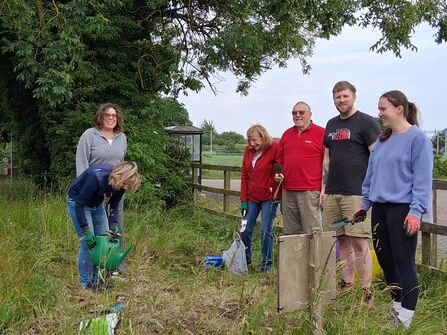 A group of six people are working outdoors in grassy area. One person is watering plants with a green watering can, and others are holding gardening tools, suggesting a community gardening or cleanup activity.
