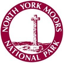 Circular logo for North York Moors National Park featuring a stone cross on a rocky base, with the park's name arced around the top and bottom in maroon lettering.