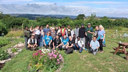 A large group of volunteers pose in a community garden beside a colourful flower bed. Raised growing plots and vegetable beds surround the group, with rolling countryside and distant hills visible in the background under a partly cloudy sky. The volunteers are standing and kneeling on the grass during a gardening session on a sunny day.