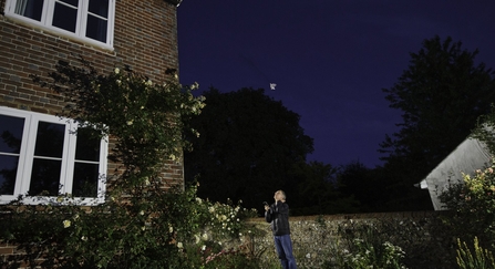 Man watches a brown long-eared bat emerge from a house roof whilst stood in his garden at night.