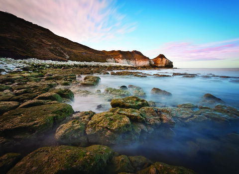 A long exposure photograph of Flamborough's rocky shore. In the foreground the sea washes over the rocks and the sky behind is a rich blue.