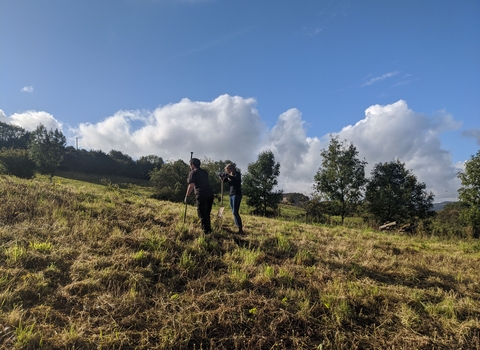 Two employees on a grassy hillside, trees in the background and blue skies with a few clouds