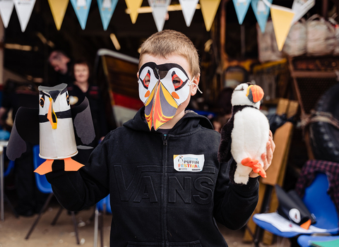 A child wearing a puffin mask holding a puffin figure he's made.