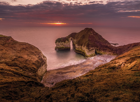 A cliff that juts out into the sea that looks like a dinosaur drinking. The sun is setting over the sea