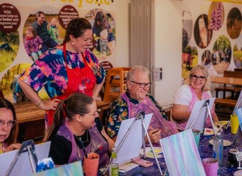 A group sat around a long table with easels in a painting class. There is a teacher standing behind admiring a student's work and everyone is looking toward's the man's work and smiling.