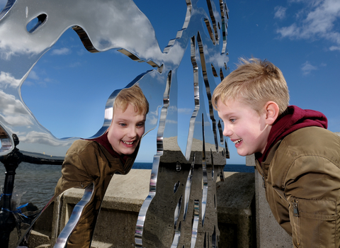 boy looking into the reflective metal of a sculpture depicting seaweed on the promenade at the coast.
