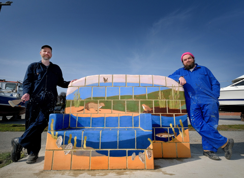 2 men stood either side of a concret bench on a seafront on a sunny day. The back of the bench is painted brickwork to depict a coastal image of the sea and cliff edges.