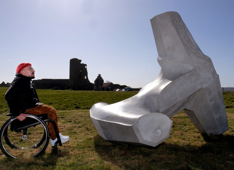 Man in wheelchair facing to the right looking at a white sculpture laid on a grassy floor on a coastal clifftop.