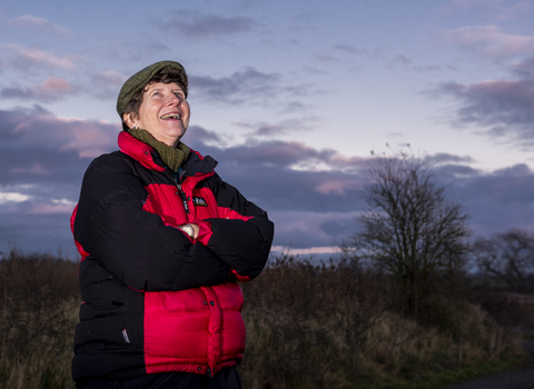 woman stood with arms folded smiling looking upto the twillight sky dressed in a coat and flat cap