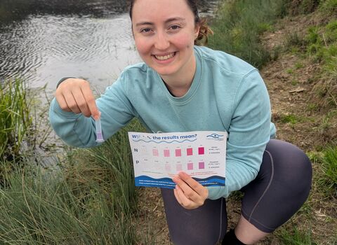 Volunteer smiles into camera, holding water quality testing kit and colour chart