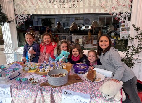 Six smiling girls pose behind a table laden with cupcakes, brownies, and a cake, a sign that says "Buy a Cake" is placed in front of the table. The girls are holding stuffed animals; a shark, and a care bear. The backdrop is a butcher shop with the sign "Bakers Butchers."