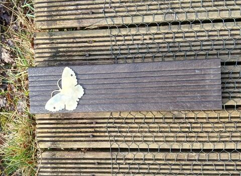 A flat wooden plaque with a white butterfly silhouette attached, set on a wooden surface with wire mesh, surrounded by grass and leaves.
