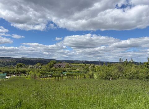 A lush green field in the foreground, with a fenced garden area and distant buildings set against a dramatic sky filled with fluffy white clouds.