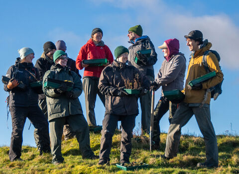 A diverse group of people wearing outdoor clothing and hats stand on a grassy slope under a bright blue sky, holding trays filled with young plants and soil.