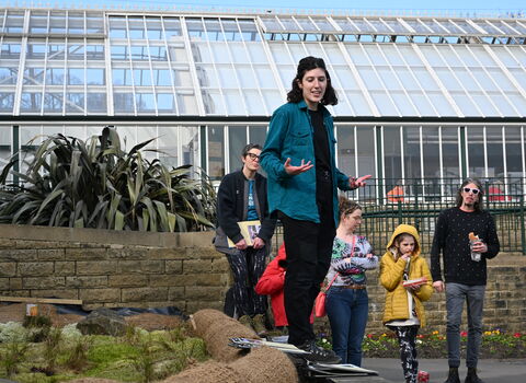 A group of people stand outdoors with a large glass greenhouse in the background; one person in the foreground, dressed in a teal shirt, is speaking with their hands gesturing.